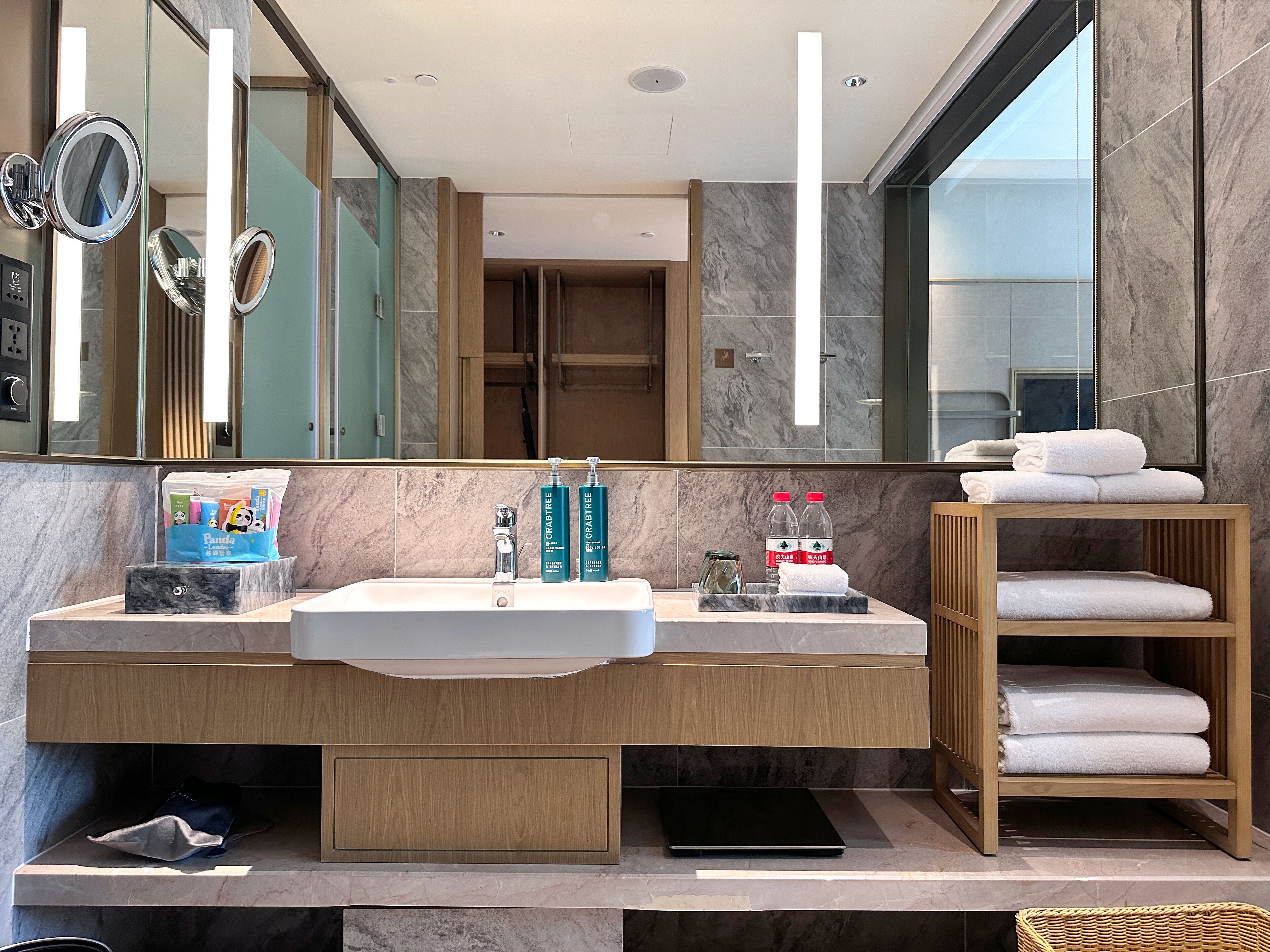 A modern bathroom with a large mirror, two sinks, and a wooden vanity. There are toiletries on the counter, and a towel rack with folded towels.