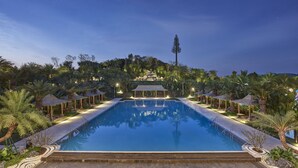 Una amplia piscina rectangular con superficie de agua azul cristalina, rodeada de palmeras y una estructura tipo pabellón.