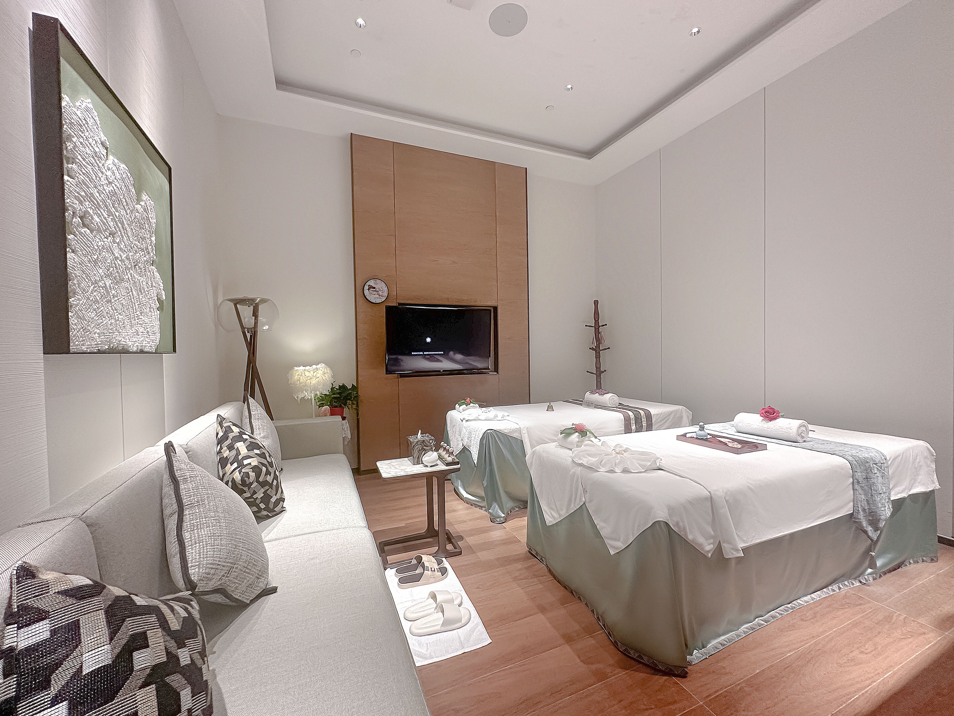 Quarto de hotel moderno com sofá, duas camas com roupa branca, televisão de tela plana, mesinha com um vaso de flores e piso de madeira.