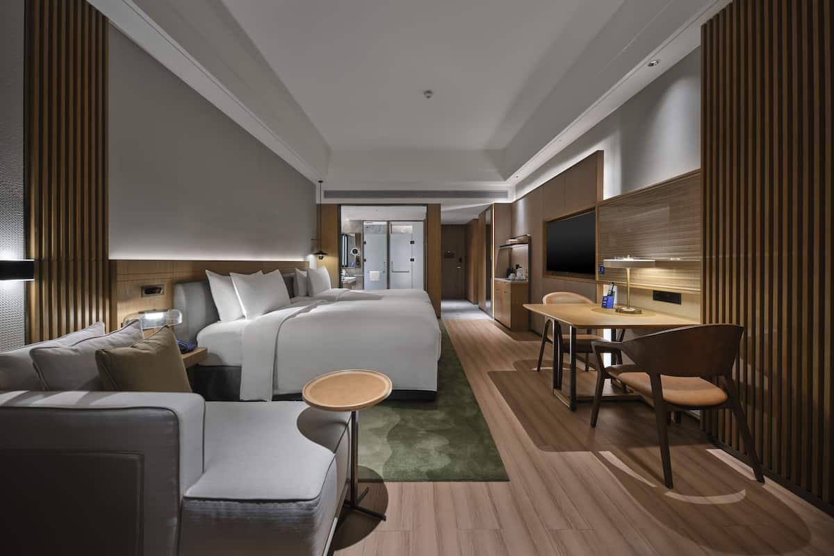 A modern hotel room with a large bed, a sofa, a small table, a TV, and a desk with a chair.