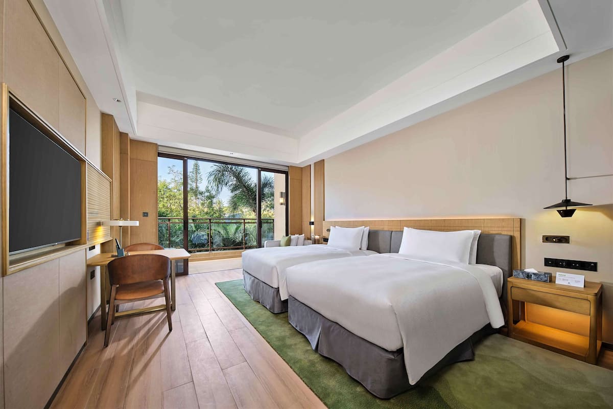 A modern hotel room with two beds, a desk, a chair, a TV, and a large window offering a view of greenery.