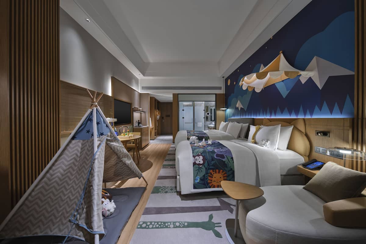 A modern hotel room with a bed, a desk, a chair, a small table, a tent, and a large wall mural.