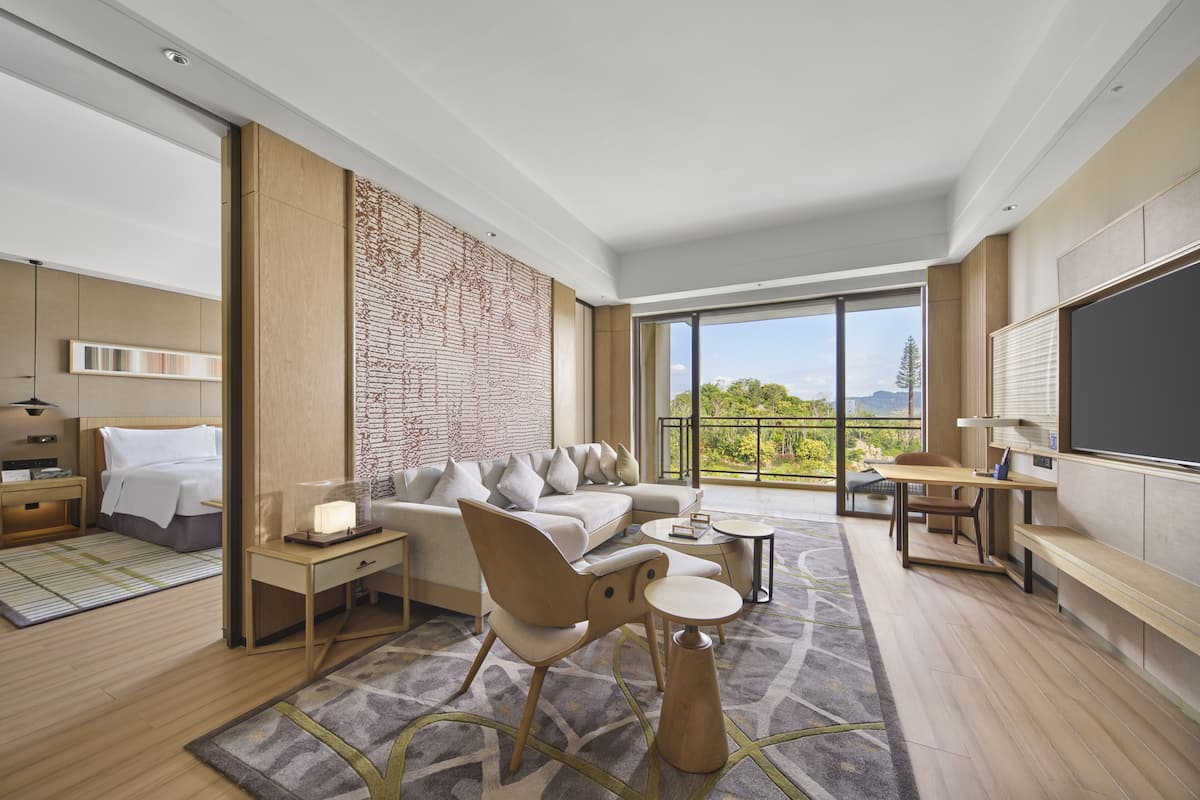 A modern hotel room with a large bed, a sofa, a desk, and a balcony with a view of the outdoors.