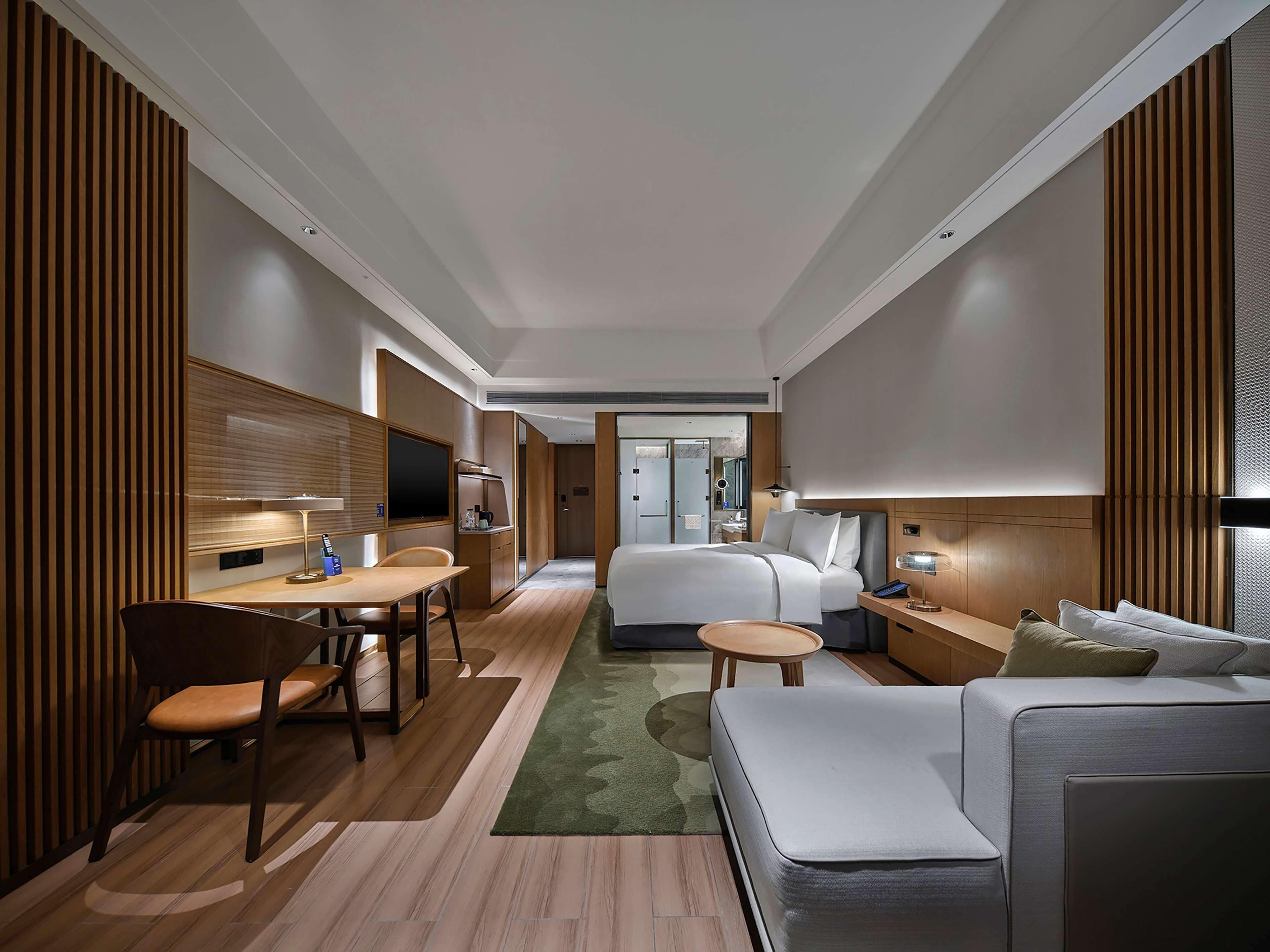 A hotel room with a bed, a sofa, a desk with a chair, a TV, and a bathroom visible through a door.