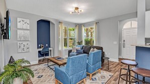 A modern living room with a sofa, chairs, a coffee table, and a TV. There are windows with curtains, a kitchen area, and a door. - 2 Bedroom Vacation Condo (2011) (Kissimmee)