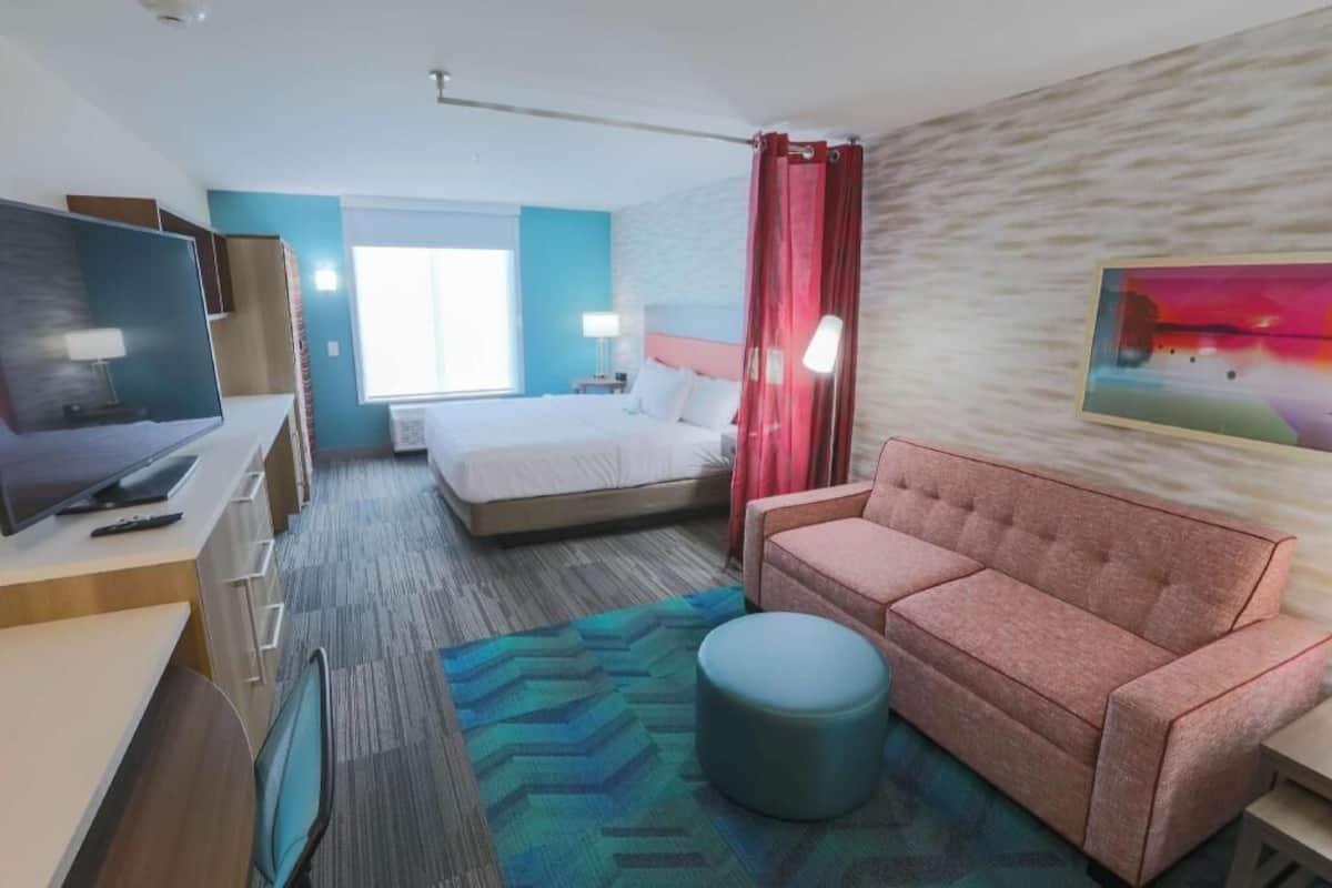 Habitación de hotel con cama, escritorio con computadora, sofá, televisor y un cuadro en la pared.