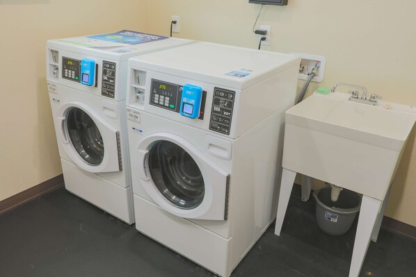 Two front-loading washing machines with control panels, a small sink, and a trash can in a utility room.