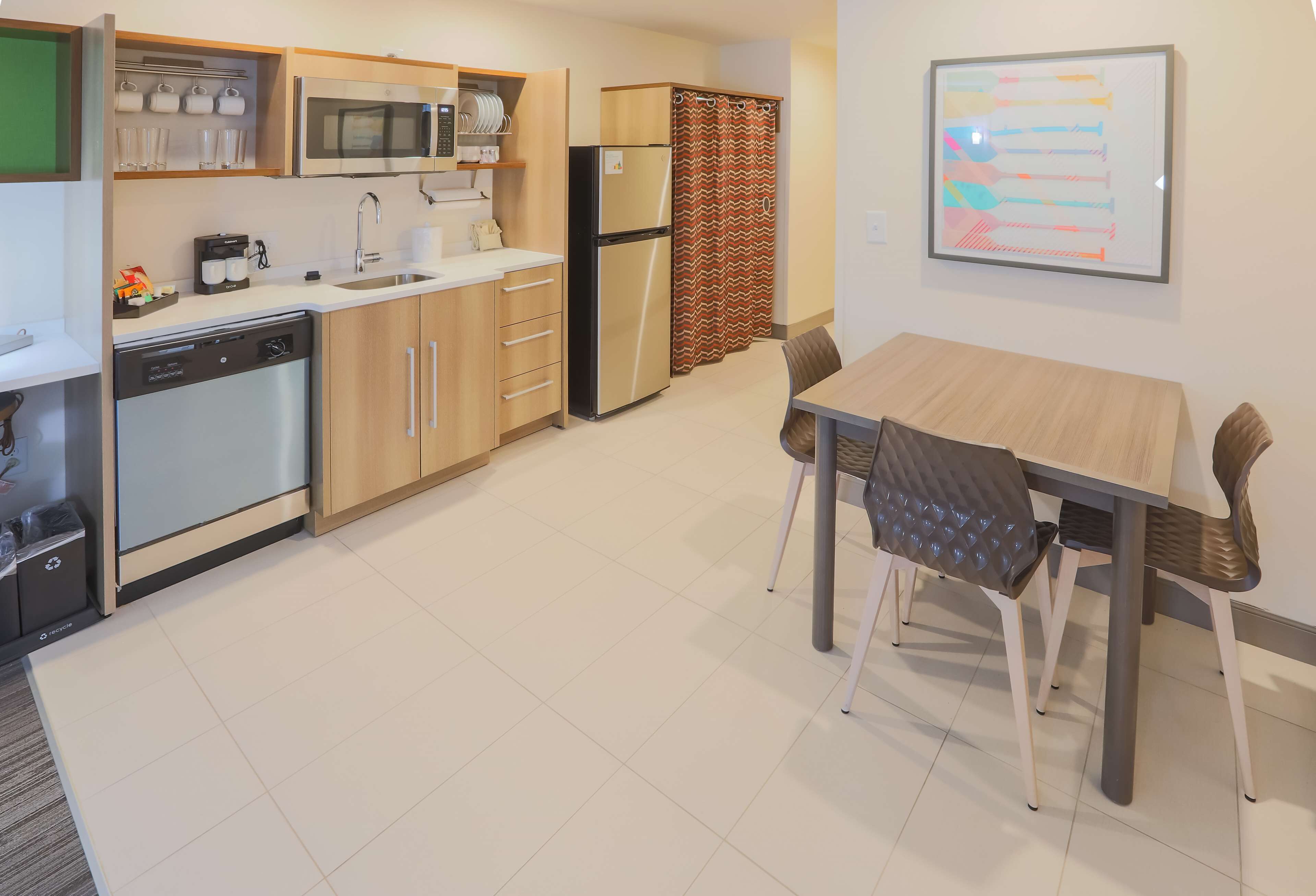 Une cuisine moderne équipée d’un four à micro-ondes, d’un réfrigérateur et d’un lave-vaisselle. On y trouve une table à manger avec des chaises, et un rideau est visible au fond.