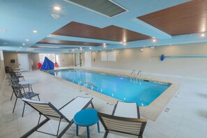 An indoor swimming pool with lounge chairs and tables, a ceiling with wooden panels, and a door leading to another room.