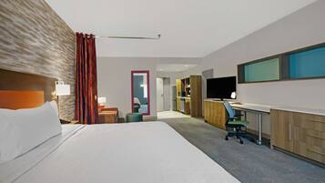 A hotel room with a large bed, a desk with a computer, a TV, and a kitchenette in the background.