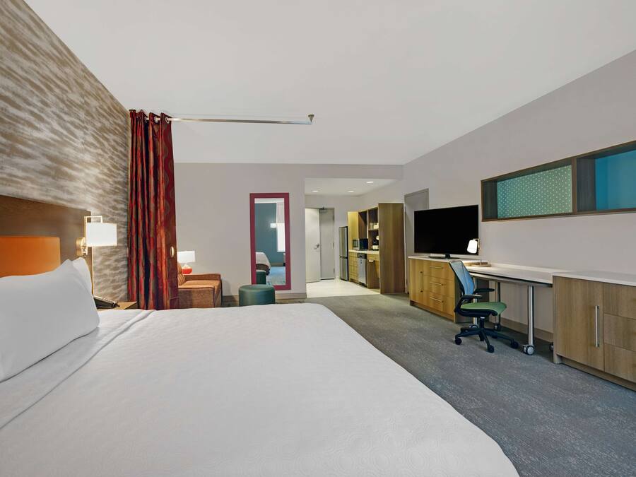 A hotel room with a large bed, a desk with a computer, a TV, and a kitchenette in the background.