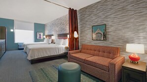 Habitació d'hotel amb un llit, un sofà, un petit otomà, una taula de nit amb una làmpada i una fotografia penjada a la paret.