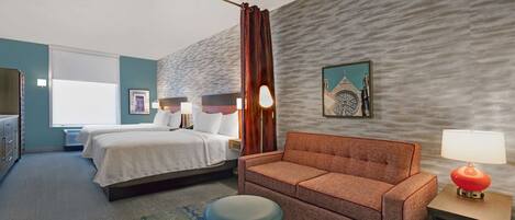 A hotel room with a bed, a sofa, a small ottoman, a nightstand with a lamp, and a wall-mounted picture.