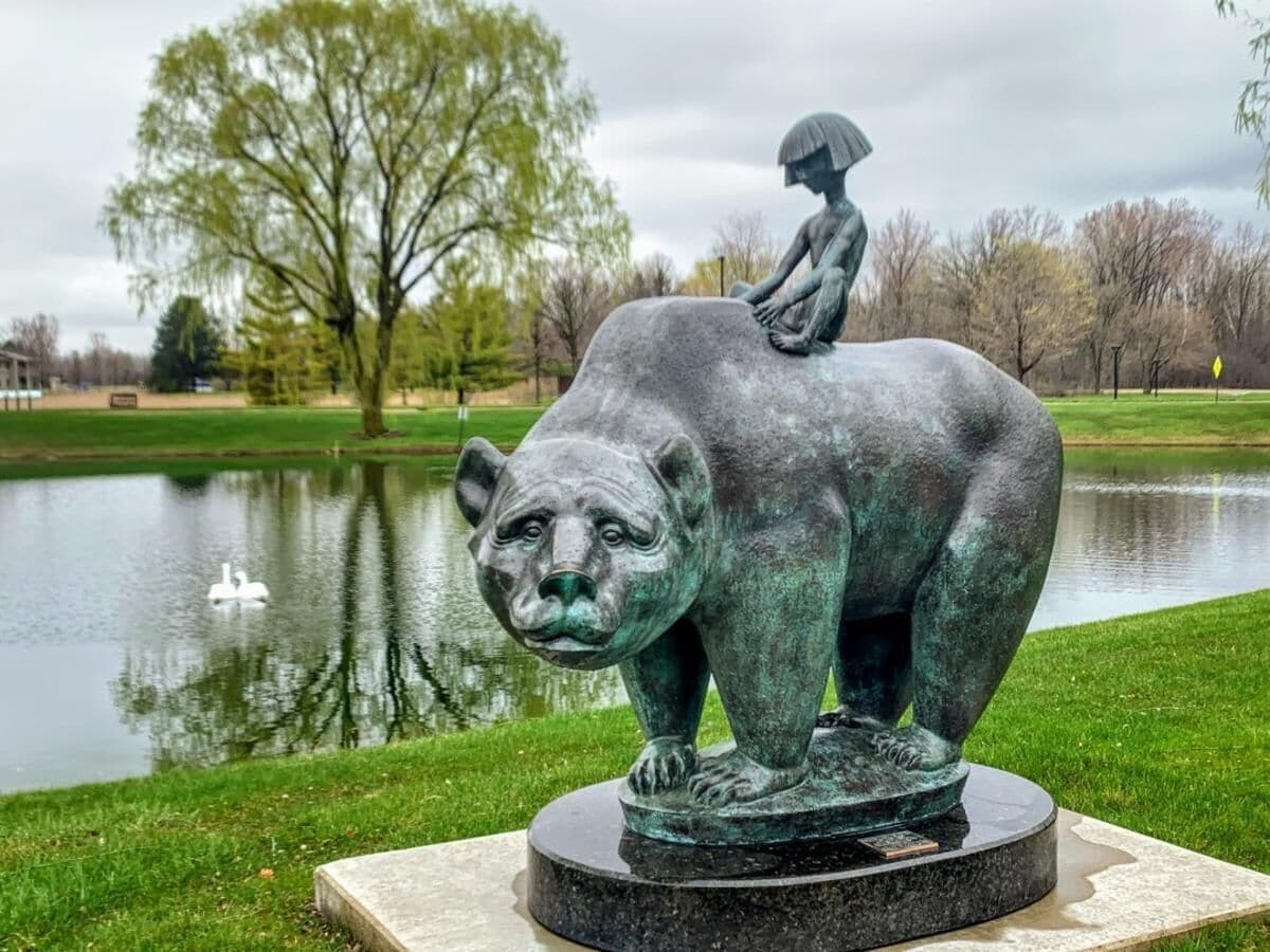 Une sculpture représentant un ours portant un enfant sur son dos, placée sur une base circulaire dans un parc où se trouvent un étang et des arbres en arrière-plan.