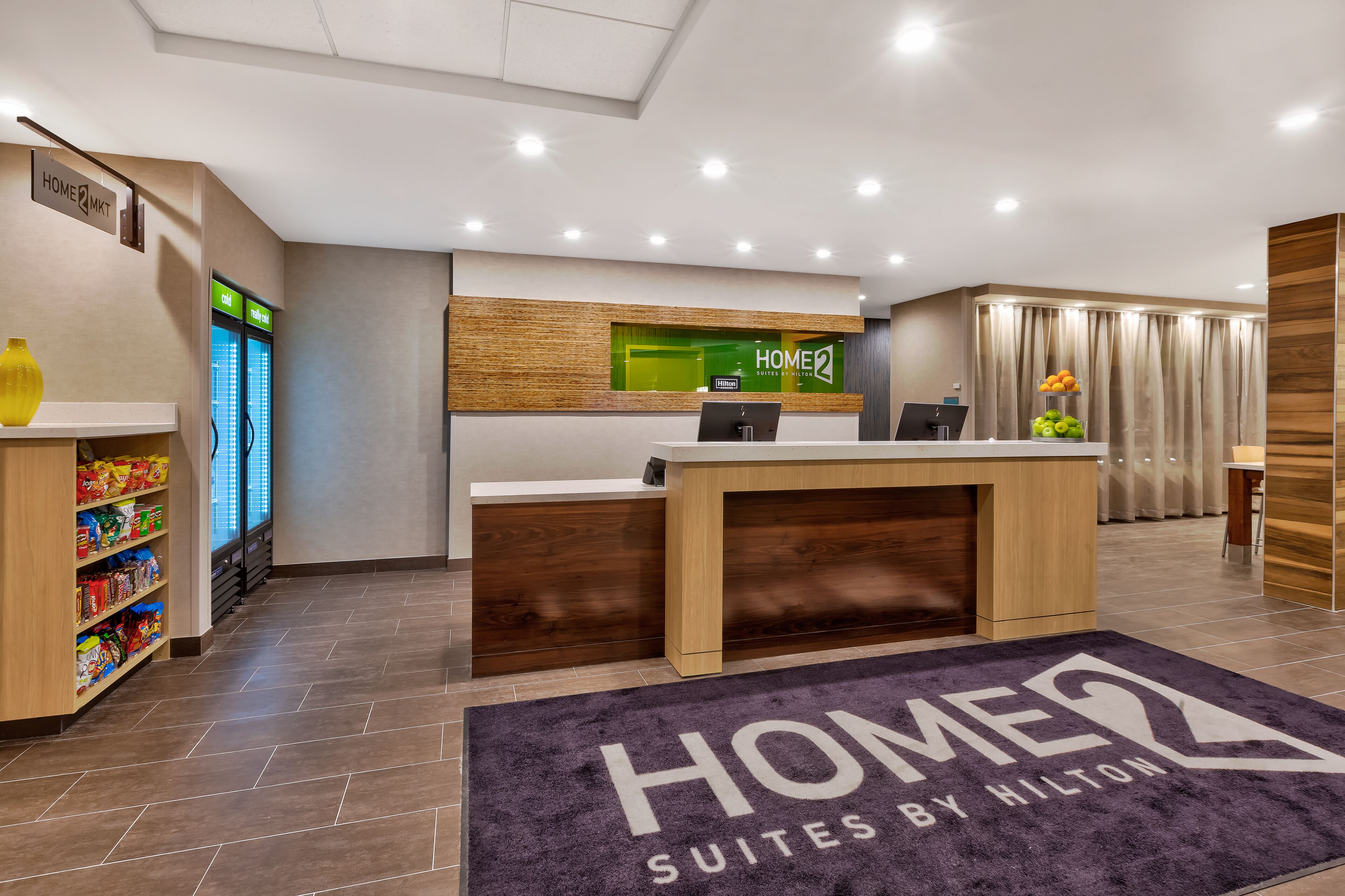 A hotel reception area with a reception desk, a digital display, a minibar, and a self-service beverage dispenser.
