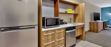 Una cocina moderna con refrigerador de acero inoxidable, microondas, lavavajillas, armarios de madera y un pequeño rincón de descanso con una silla y una mesa.