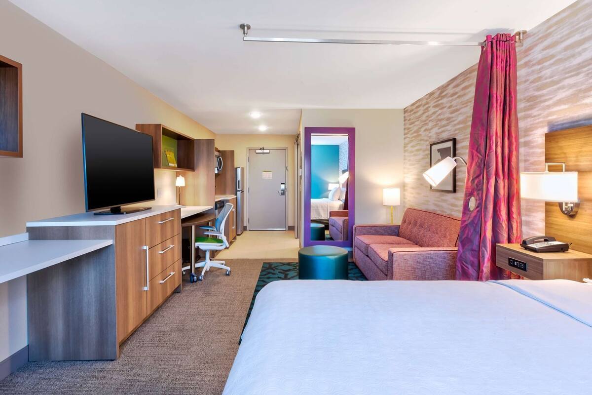 A hotel room with a bed, a desk with a TV, a sofa, a bathroom with a mirror, and a nightstand with a lamp.