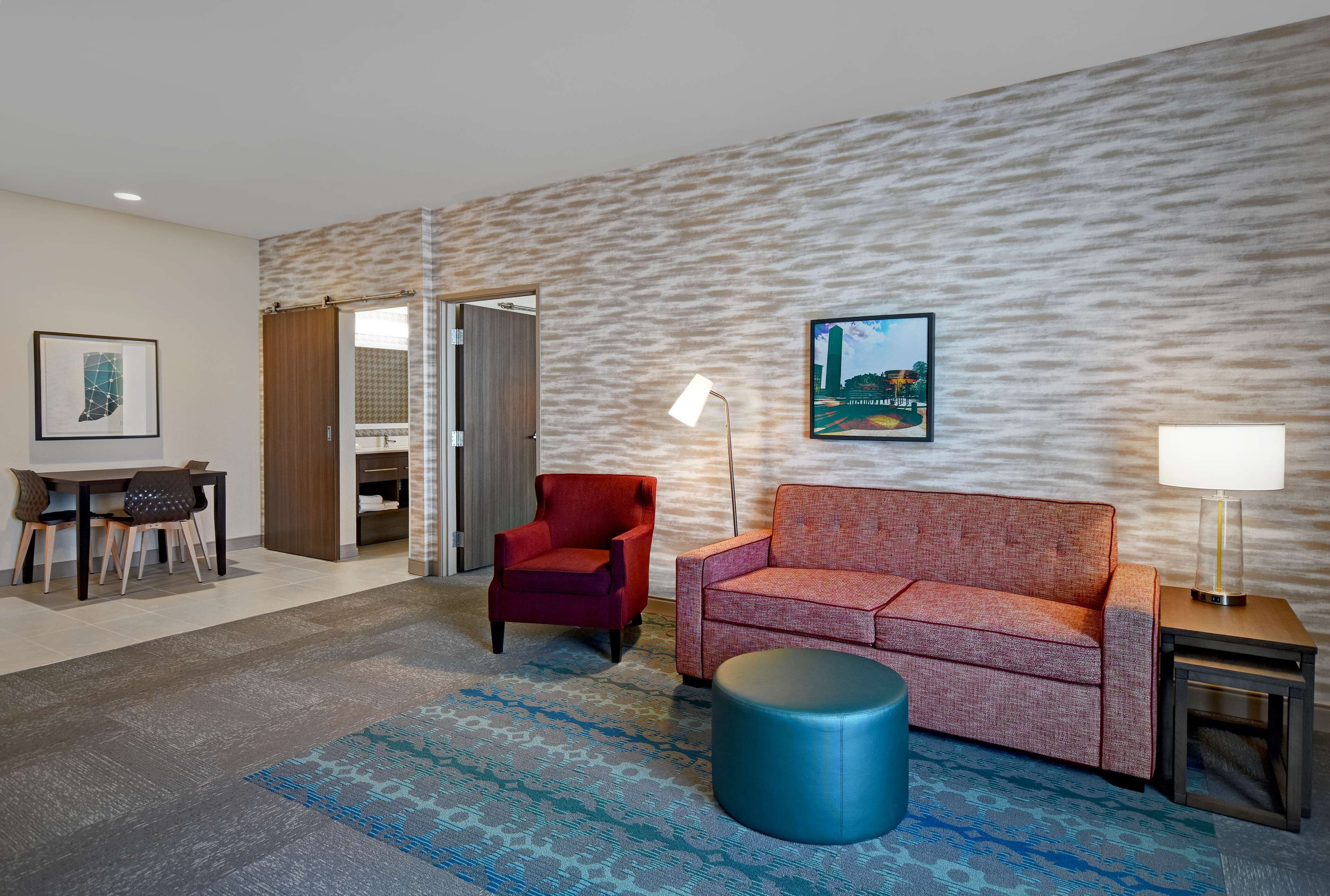 A modern hotel room with a patterned wall, a sofa, a red armchair, a round ottoman, a desk with chairs, and a framed picture on the wall.