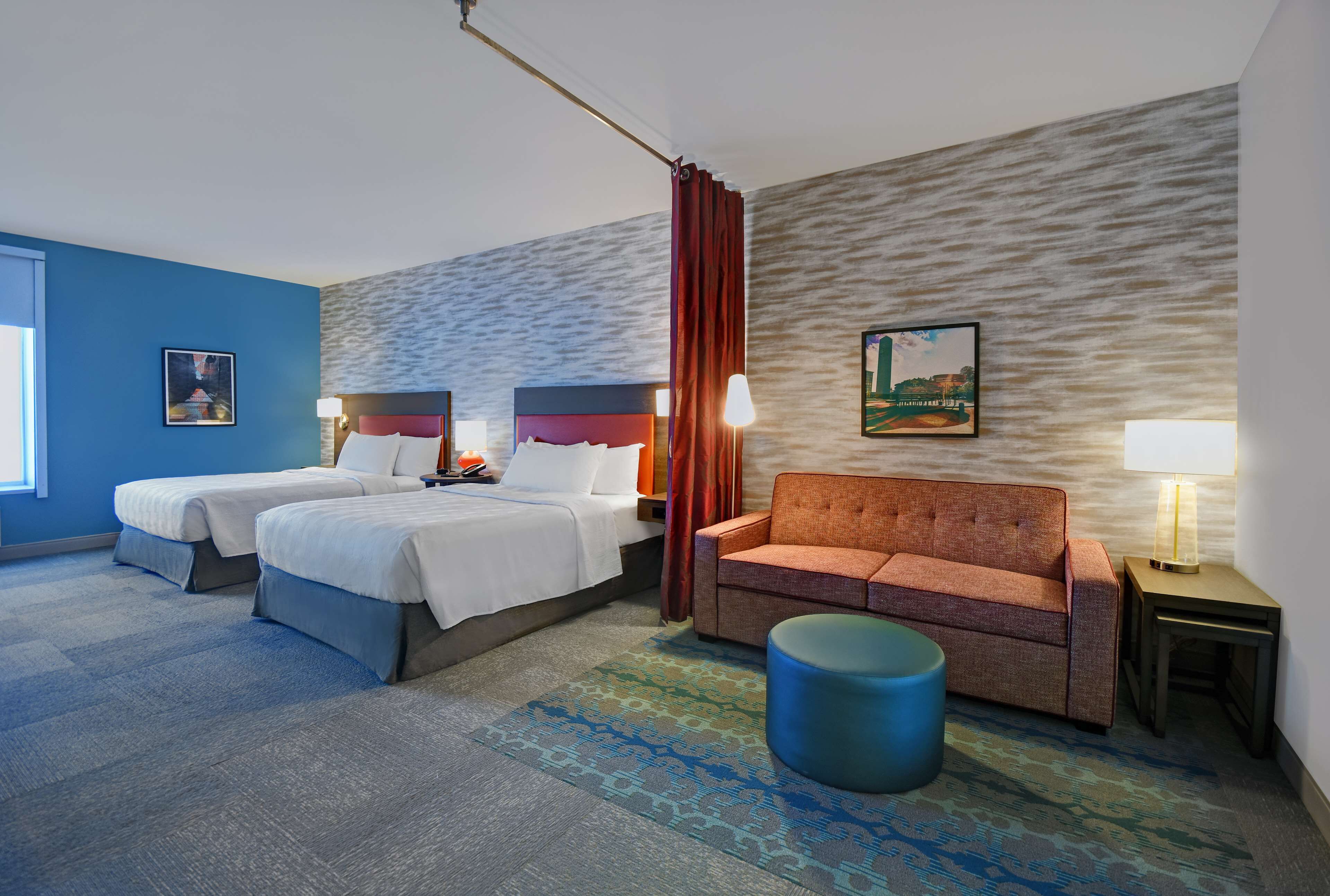 A hotel room with two beds, a sofa, a blue ottoman, and a patterned rug.