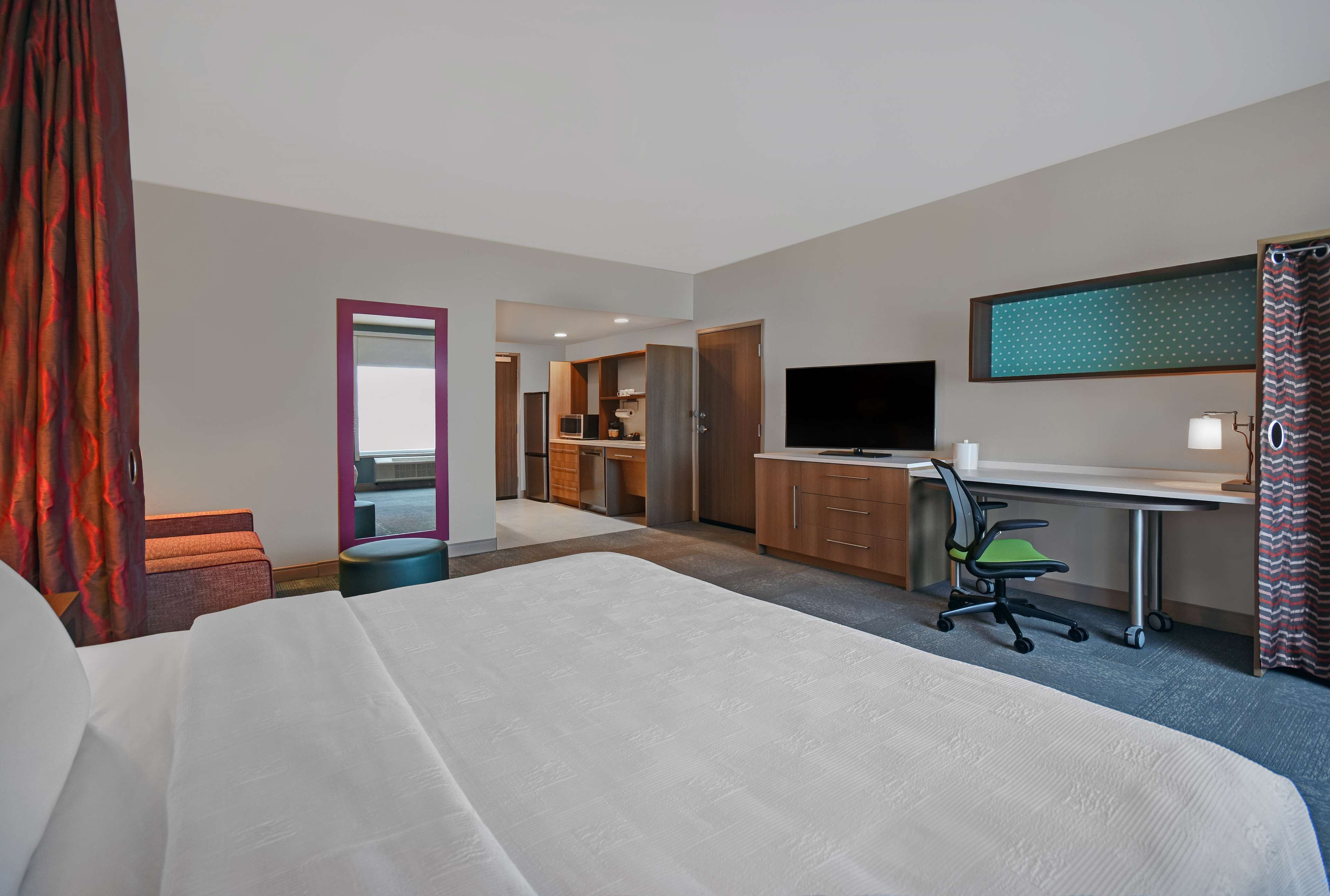A hotel room with a large bed, a desk with a computer, a TV, and a kitchenette area.
