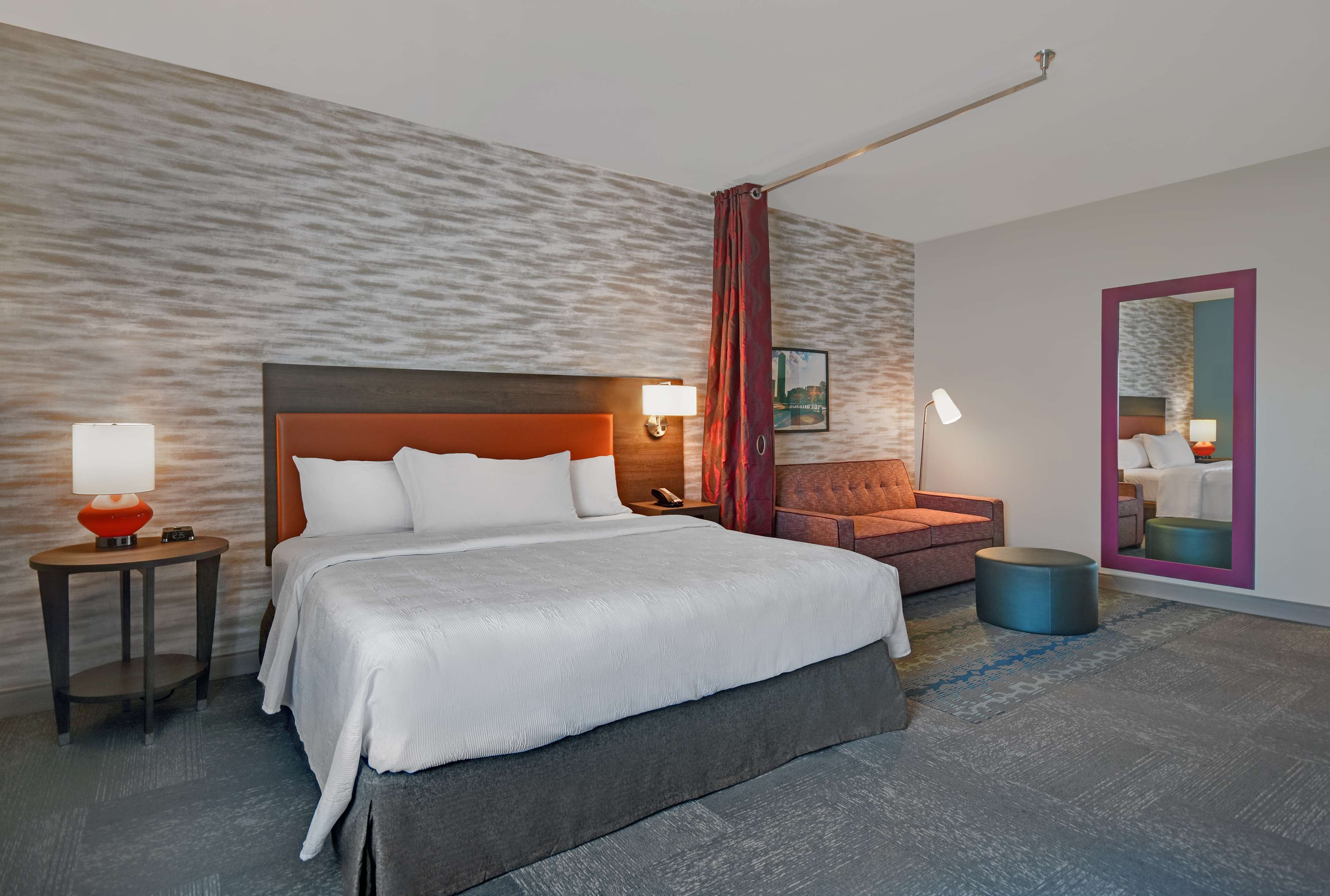 A modern hotel room with a large bed, a nightstand with a lamp, a red accent wall, and a round ottoman.