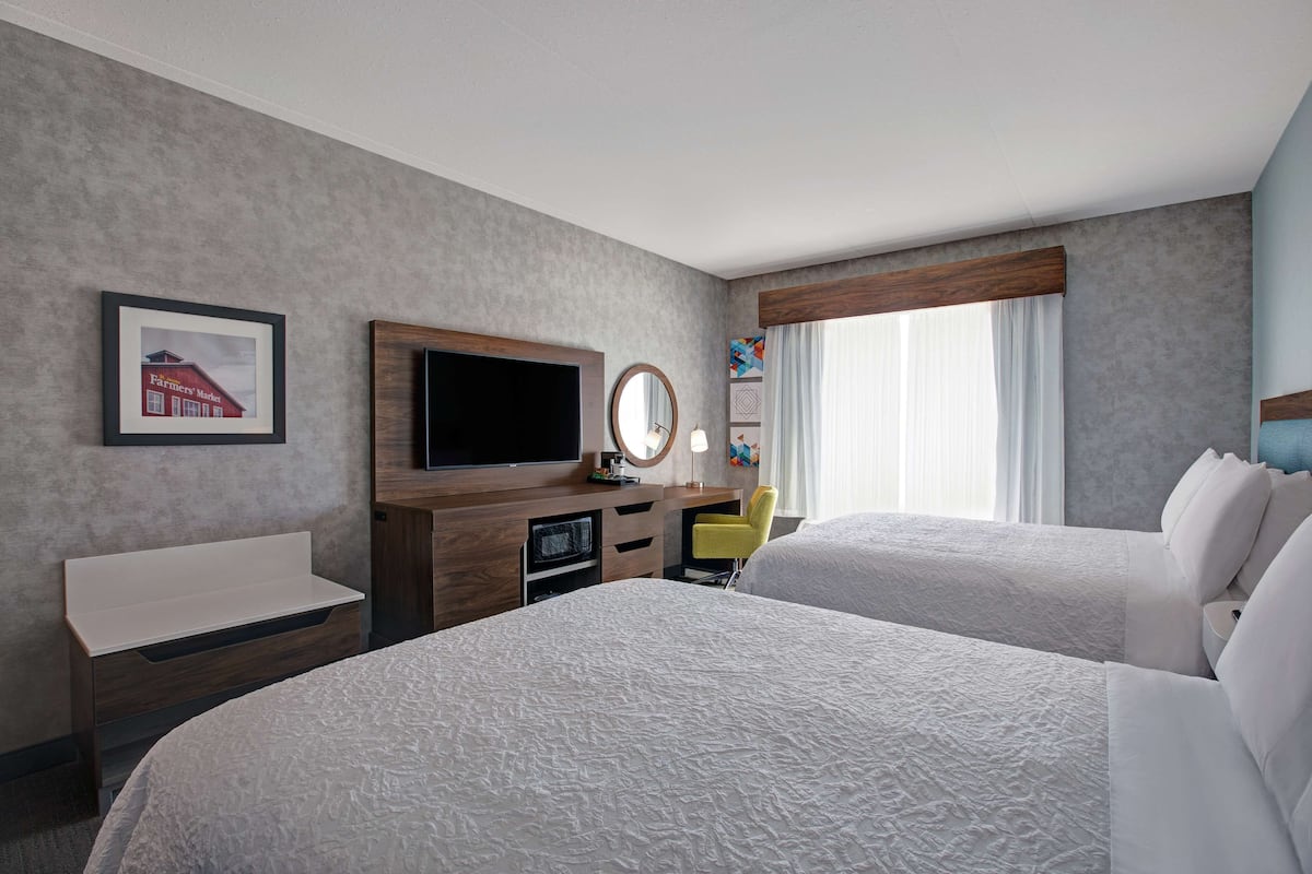 A hotel room with two beds, a TV, a desk with a chair, a mirror, and a window with curtains.