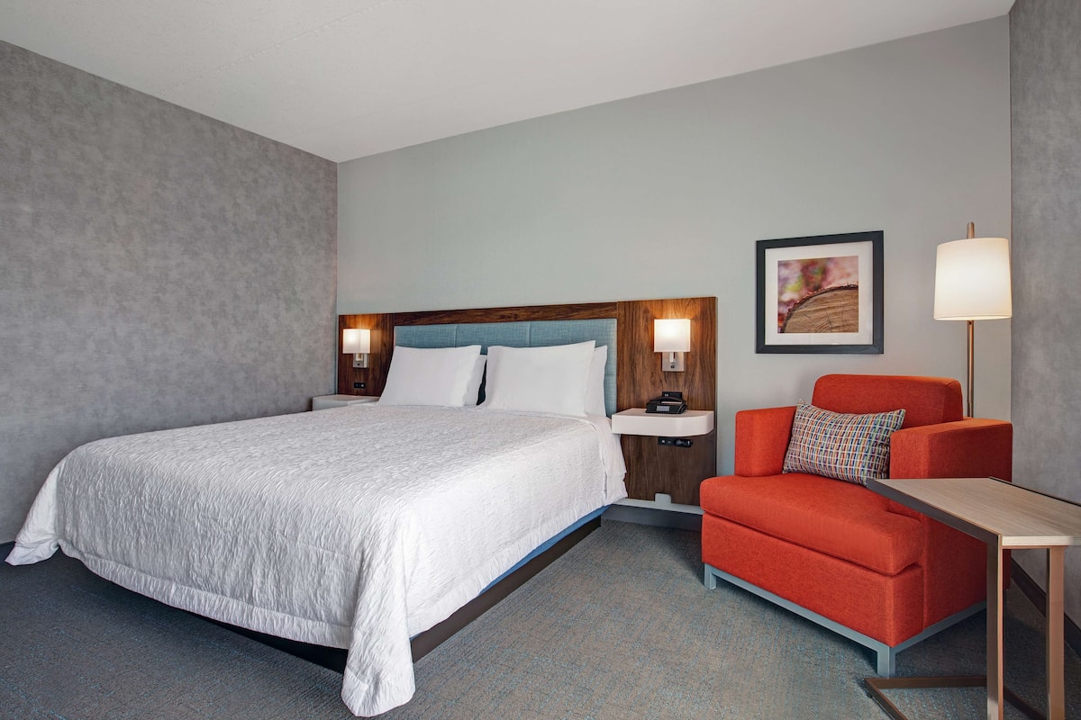A hotel room with a large bed, a red armchair, a small table, and a nightstand with lamps.