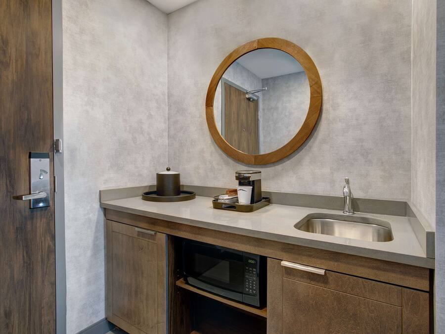 Un baño moderno con puerta de madera, un espejo redondo, un lavamanos, una cafetera y un microondas.