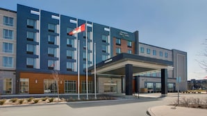 A modern hotel with a prominent entrance, a flag, and a sign indicating it is a Hampton Inn & Suites. - Hampton Inn & Suites by Hilton Waterloo St. Jacobs (Township of Woolwich)