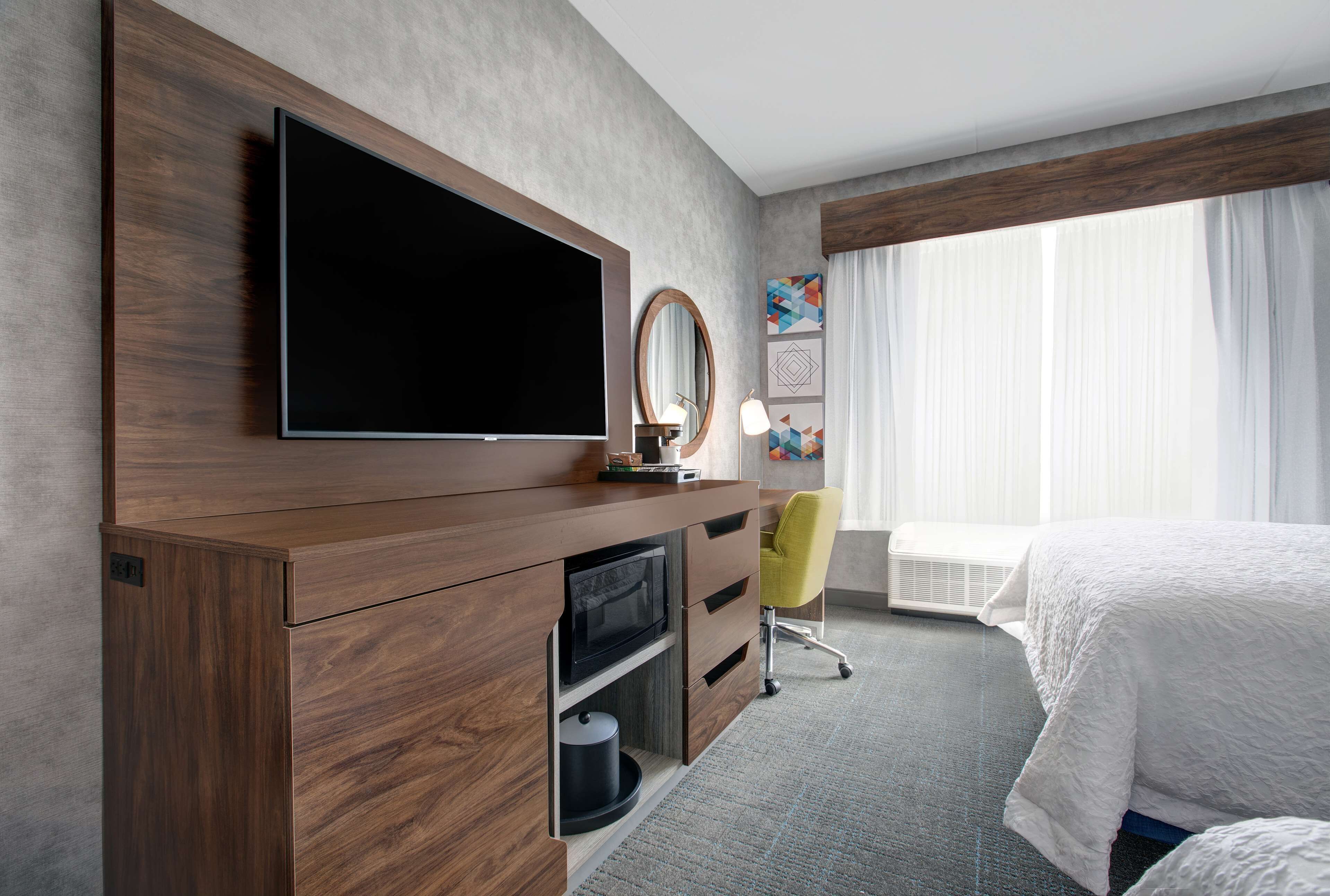 A hotel room with a large flat-screen TV, a wooden dresser, a desk with a chair, and a bed with white bedding.