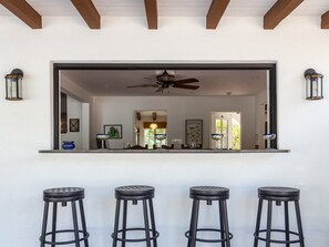 A bar area with three stools, a ceiling fan, and a view into a room with a door and artwork.