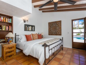 A bedroom with a bed, a bookshelf, a painting, and a view of a swimming pool.