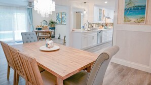 Un coin repas avec une table en bois, des chaises et un lustre. Au fond, on aperçoit une cuisine avec des meubles blancs et un tableau représentant une scène de plage.