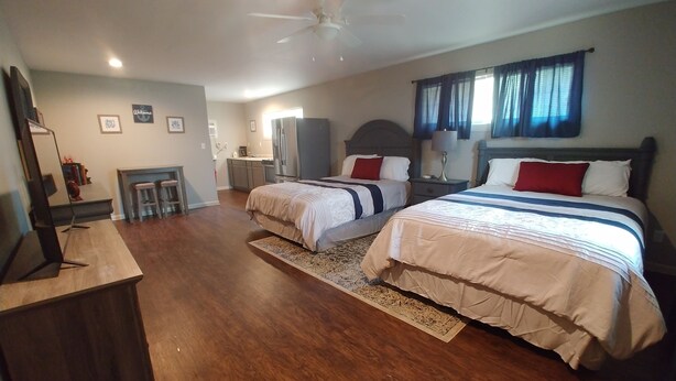 A bedroom with two beds, a desk, a chair, a ceiling fan, and a small kitchenette.