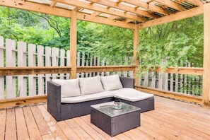 Un patio esterno con un divano in vimini e un tavolo con piano in vetro, circondato da un pavimento in legno e una recinzione di legno bianca.