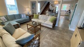 A spacious living room with a sofa, a coffee table, and a wicker chair. A staircase leads to a kitchen area.