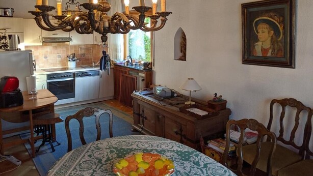A dining area with a chandelier, wooden chairs, a table with a patterned tablecloth, and a painting on the wall.