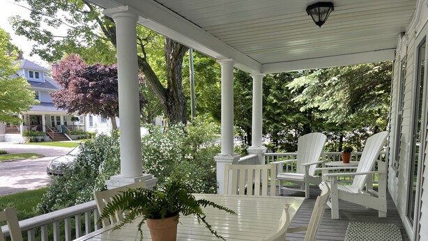 A covered porch with a table and chairs, surrounded by greenery and a house in the background. - CENTRAL A/C!!! Huge Downtown Historic CVX Home! Walk to town & beach! (Charlevoix)