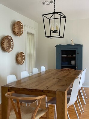 Uma sala de jantar com mesa de madeira e cadeiras brancas, um armário azul com prateleiras e arte decorativa em tecido na parede.
