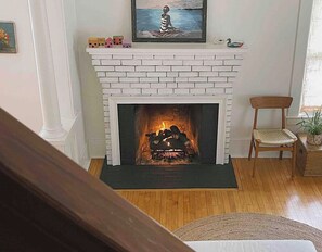 Lareira de tijolos brancos com fogo, um quadro emoldurado acima e uma cadeira de madeira ao lado.