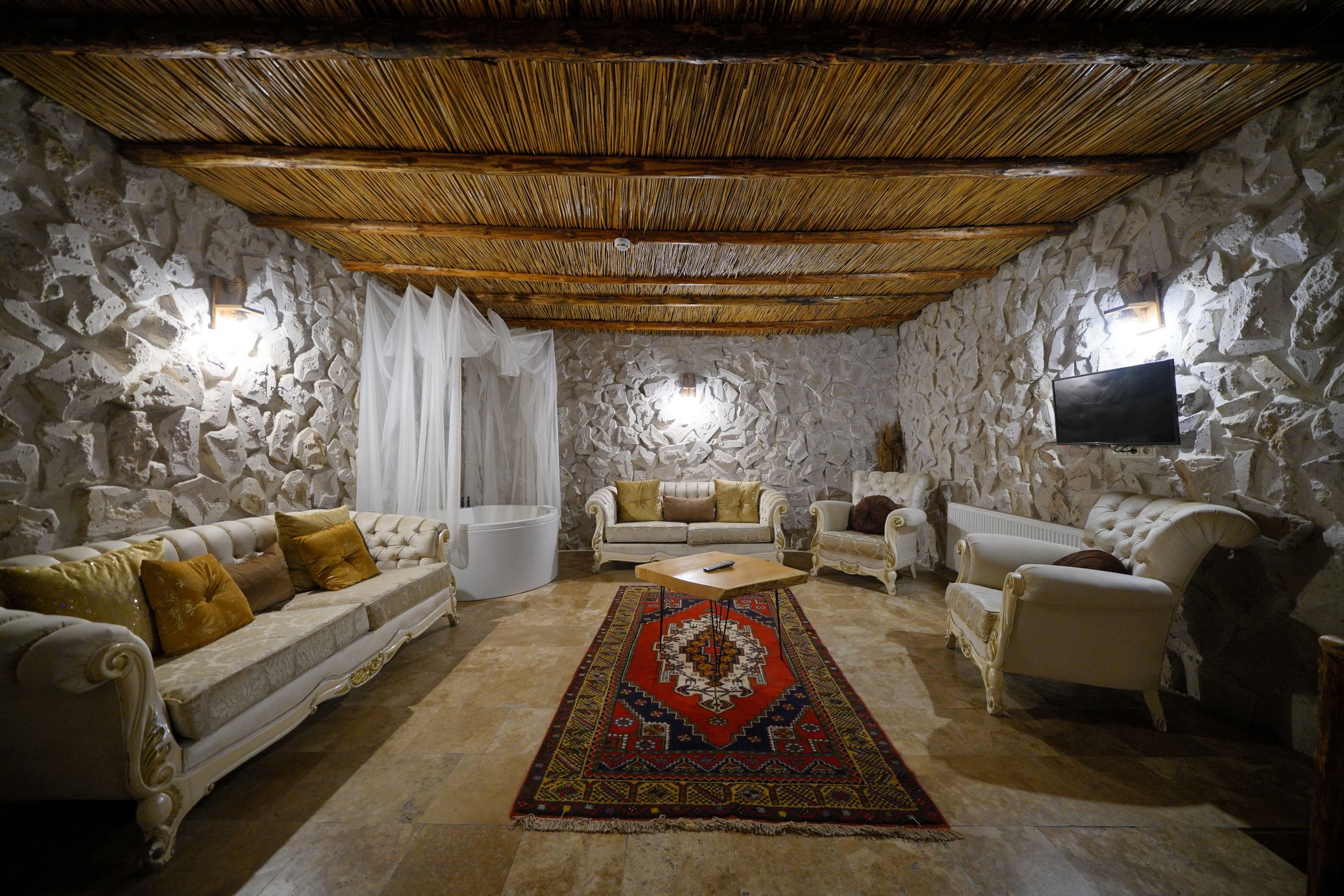 Una habitación con paredes de piedra, techo de madera, un sofá blanco, un sofá beige, un sillón blanco, un tapete con diseño rojo y negro, y un televisor de pantalla plana montado en la pared.