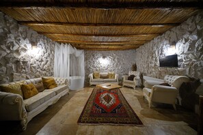 Habitació amb parets de pedra, sostre de fusta, un sofà blanc, un sofà beix, un butacó blanc, una catifa amb dibuixos vermells i negres, i una televisió plana penjada a la paret.