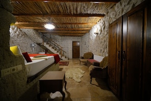Una habitación rústica con pared de piedra, techo de madera, una cama con manta roja, una mesa de madera, una silla y un armario de madera.