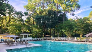 Piscina retangular com espreguiçadeiras e guarda-sóis, cercada por árvores e uma cerca branca.