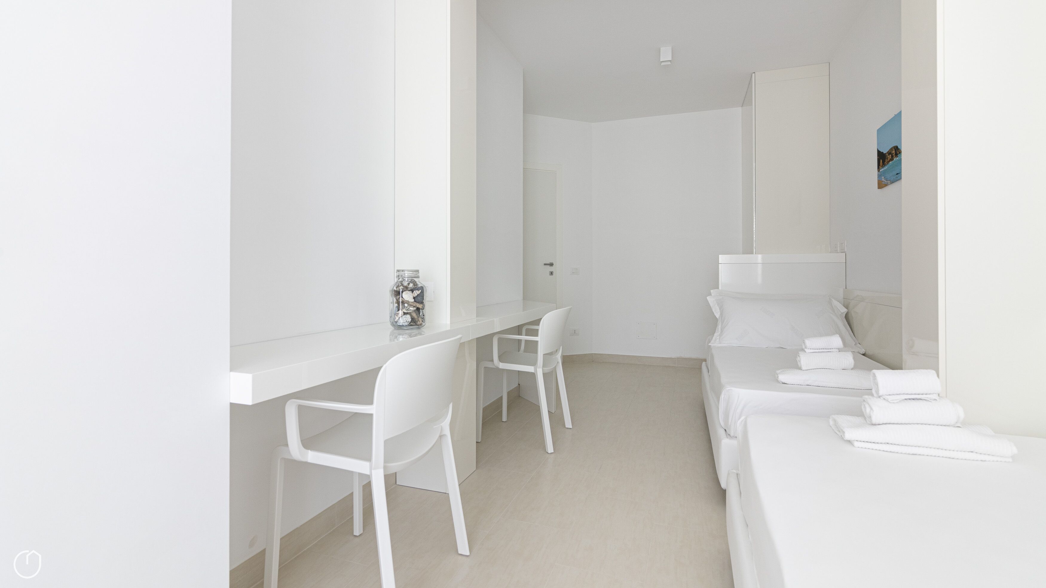 Habitació blanca i minimalista amb escriptori, dues cadires, un llit i tovalloles ordenadament dobles, i un petit quadre a la paret.