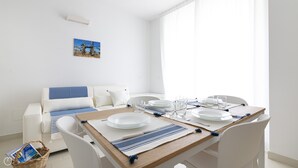 Un comedor con una mesa de madera dispuesta para cuatro personas, sillas blancas y un sofá blanco con cojines azules. Un cesto con revistas está en el suelo.