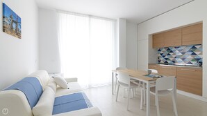 Una sala de estar moderna con sofá, mesa de comedor y sillas. Se observa una zona de cocina con salpicadero de azulejos y un lavabo.