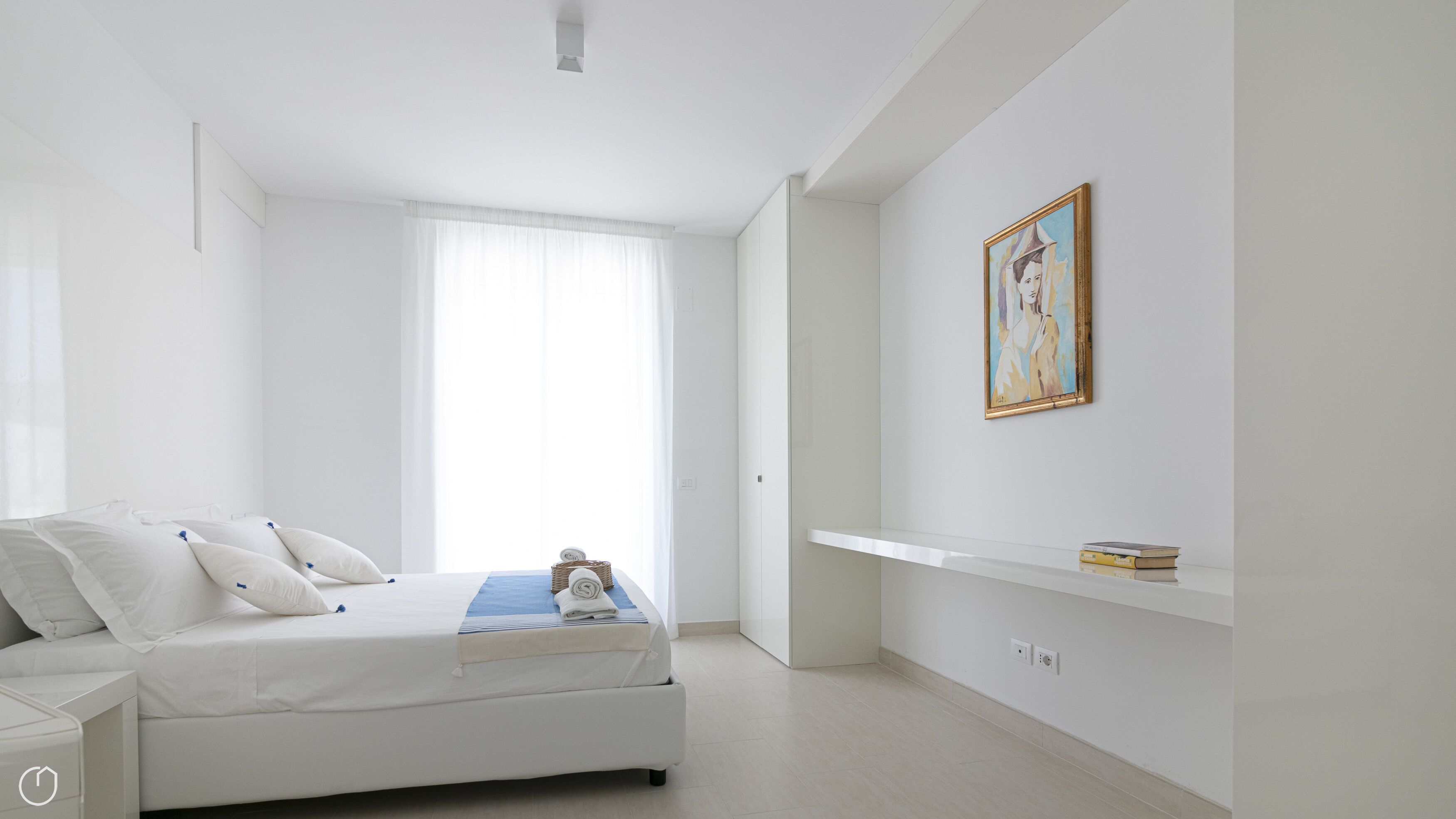 Un dormitorio minimalista con cama, mesita de noche, ventana con cortinas translúcidas, estante de pared con libros y un cuadro enmarcado.