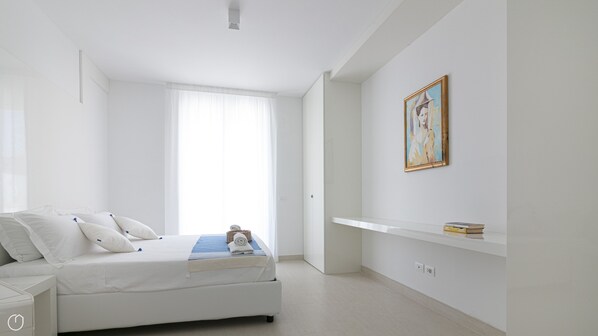 A minimalist bedroom with a bed, a bedside table, a window with sheer curtains, a wall-mounted shelf with books, and a framed painting. - Rivazzurra Homes - 41 (Termoli)