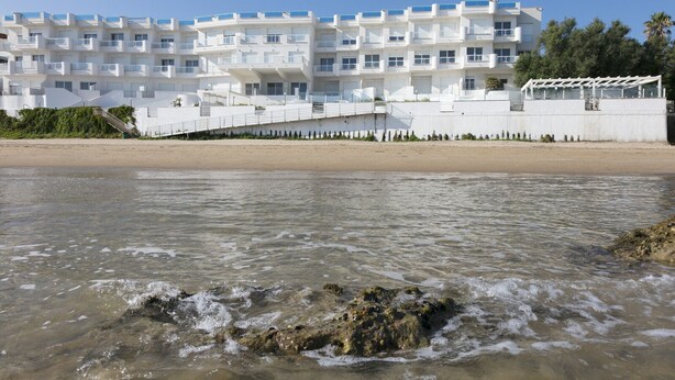 Un hotel frente a la playa con fachada blanca, varios balcones y vista despejada al mar.